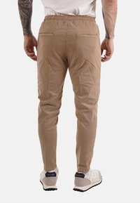 Uomo in pantaloni beige slim-fit e sneakers bianche, in piedi con le mani rilassate, tatuaggi visibili sulle braccia, sfondo bianco.