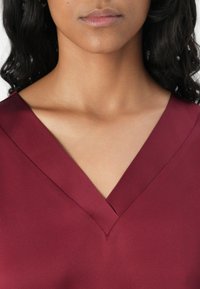 Blouse bordeaux avec un décolleté en V, tissu lisse et design structuré, avec un léger éclat et sans embellissements visibles.