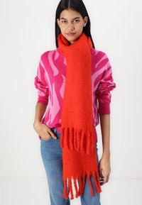 Bufanda roja y esponjosa con bordes flecados, llevada sobre un suéter rosa con estampado. La textura suave y el color vibrante realzan el atractivo visual.