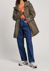 Oliwkowa parka z kapturem i pomarańczowym podszyciem, noszona na koralową bluzkę i niebieskie dżinsy. Buty to białe i złote trampki z sznurowadłami.