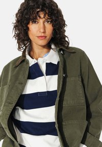 Veste verte olive avec un col, dotée de deux poches avant. En dessous, un polo rayé bleu marine et blanc.