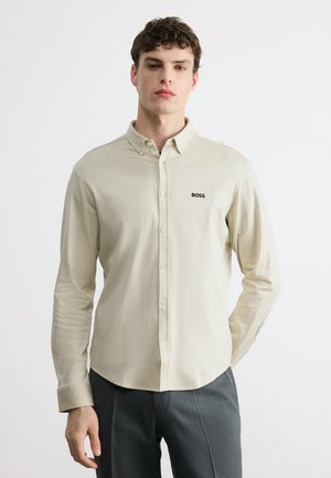 Jeune homme portant une chemise beige à boutons avec le logo "BOSS" sur la poitrine et un pantalon gris foncé, debout devant un fond blanc uni.