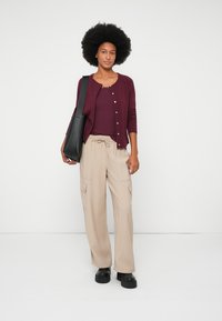 Cardigan burdeos con botones sobre una blusa ajustada de canalé, combinado con pantalones cargo beige. La modelo lleva un bolso negro de hombro y zapatos negros de plataforma.