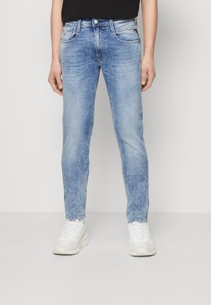 Man wearing slim-fit light blue washed jeans, white sneakers, and a black shirt standing against a neutral background.