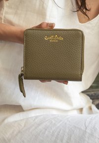 Alyvuogių žalios odos piniginė su tekstūruota paviršiumi, auksinėmis iškaltais ženklu ir užtrauktuku, kuriame yra mažas odinis užtrauktuko traukiklis.