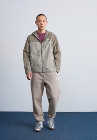 Veste zippée légère pour homme, de couleur vert olive avec des panneaux gris, associée à un pantalon de jogging beige et des chaussures de sport argentées. Fond uni.