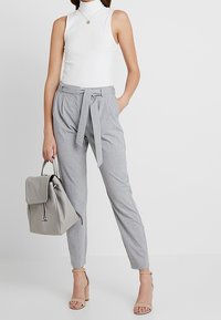 Světle šedé kalhoty s jemným proužkem a zavázaným pasem, doplněné přiléhavým bílým žebrovaným topem a svetle šedým batohem.