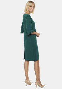 Turkusowa sukienka do kolan z falbaniastymi rękawami o długości trzech kwart, bocznym rozcięciem i gładką, jedwabistą fakturą. Noszona z beżowymi szpilkami.