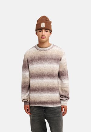 Junger Mann mit brauner Beanie und gestreiftem Strickpullover in Beige-, Braun- und Grautönen, vor einfarbigem Hintergrund stehend.