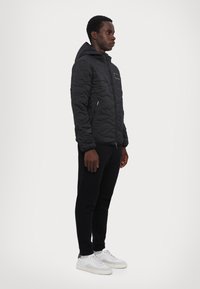 Sort quiltet jakke med hætte, med en frontlynlås og sidelommer, parret med sorte bukser og hvide sneakers.