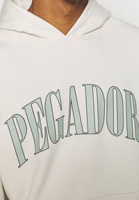 Felpa con cappuccio beige dalla texture morbida, con una grande scritta "PEGADOR" in verde chiaro sul petto. Cappuccio e dettagli di cuciture sottili visibili.