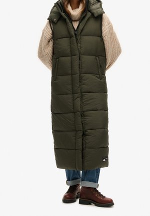Frau trägt ärmellosen langen olivgrünen gesteppter Mantel, beigen grob gestrickten Pullover, hochgekrempelte Jeans und braune Lederstiefel.