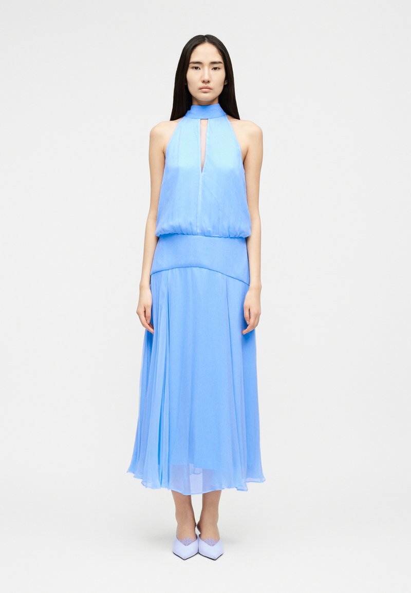 Νεαρή γυναίκα με μακριά σκούρα μαλλιά που φοράει αμάνικο ανοιχτό μπλε φόρεμα με σχέδιο halter και ταιριαστά τακούνια, στέκεται μπροστά σε λευκό, απλό φόντο.