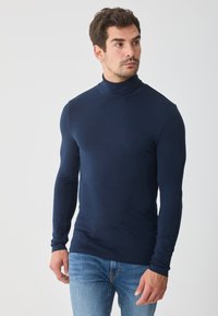 Tricou cu guler înalt, de culoare albastru marin, cu mânecă lungă, realizat dintr-un material moale, cu un design ajustat și o textură netedă, asortat cu blugi de culoare albastru deschis.