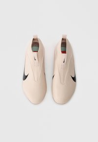 Gaiši persiku slip-on apavi, kas izgatavoti no teksturēta materiāla, ar melnu Nike swoosh, minimālistisku dizainu un vilkšanas cilpu uz papēža.