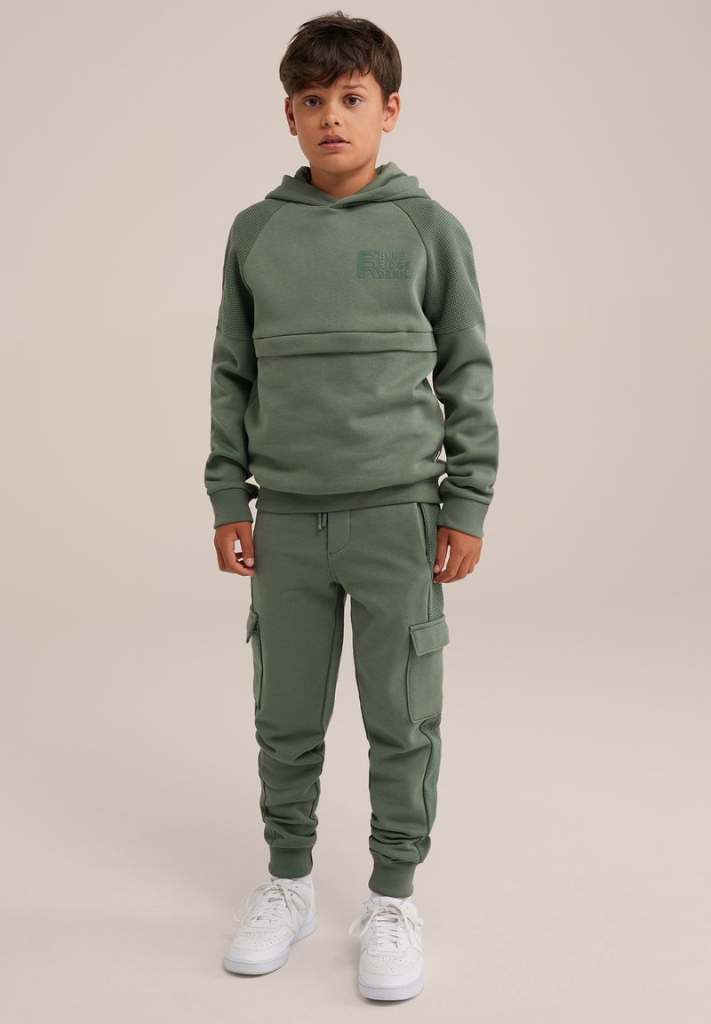 Zaļa sporta džempera s kapuci un logotipu kombinācija ar atbilstošām kabatas biksēm, kurām ir sānu kabatas. Baltas kedas noslēdz tēlu.