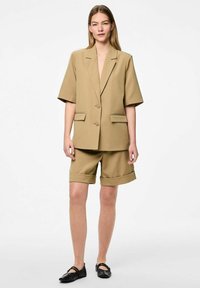 Blazer oversize en tan avec des manches courtes, deux poches avant et deux boutons, associé à un short roulé assorti. Porté avec des chaussures noires plates.