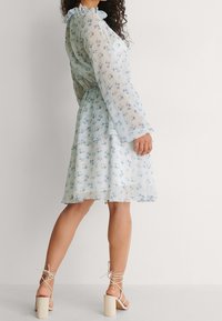 Robe florale bleu clair avec de longues manches évasées et un col volanté. Fabriquée en tissu transparent avec un design en niveaux et des sandales assorties.