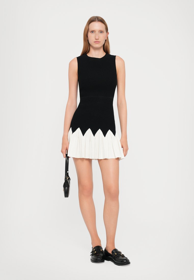 Robe noire sans manches avec un corsage ajusté et une jupe plissée blanche présentant un ourlet en zigzag. Le mannequin tient un sac à bandoulière noir et porte des chaussures noires.