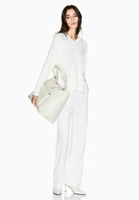Weißer, gestrickter V-Ausschnitt-Pullover, kombiniert mit passenden, weit geschnittenen Hosen. Das Model hält eine weiche, cremefarbene Ledertasche mit klarer Struktur.