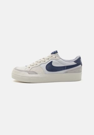 Biały niski sneaker z ciemnoniebieskim logo Nike, z przodu sznurowany i grubą, kremową gumową podeszwą na jednolitym tle.