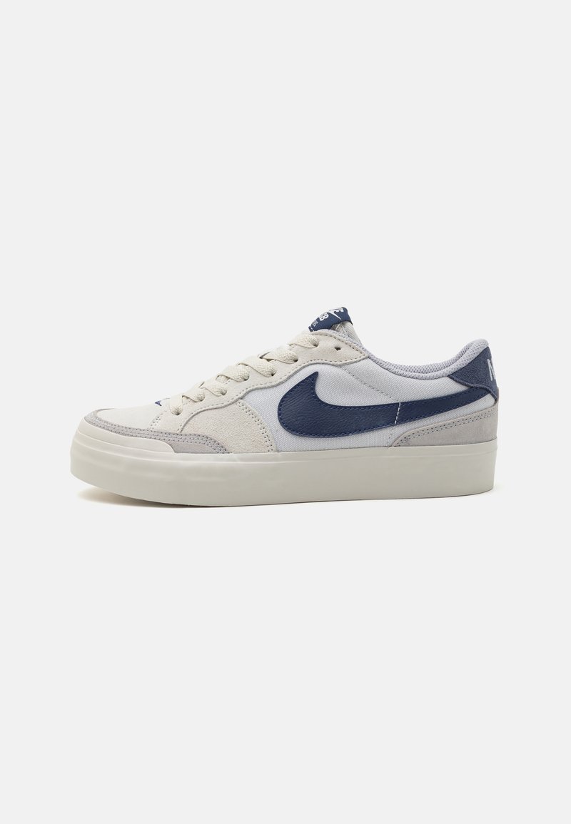 Baskets basses blanches avec logo Nike bleu foncé, laçage à l'avant et semelle épaisse en caoutchouc blanc cassé sur fond uni.