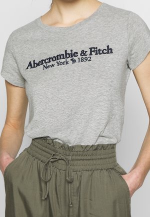 Kvinde iført en grå Abercrombie & Fitch t-shirt og grønne bukser med høj talje og snørebånd, hænderne i lommerne.
