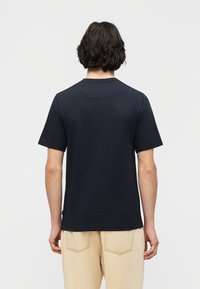 Άτομο με μεσαίου μήκους σκουρόχρωμα μαλλιά που φοράει απλό μπλε σκούρο κοντομάνικο πουκάμισο και ανοιχτό μπεζ παντελόνι, με την πλάτη γυρισμένη, σε λευκό φόντο.