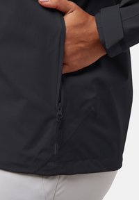 Giacca nera impermeabile con tasca laterale a zip, texture liscia e polsini regolabili. Design minimalist senza loghi o fantasie visibili.
