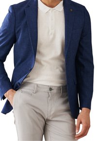 Granatowa teksturowana marynarka o dopasowanym kroju, noszona na jasnobeżowej koszulce polo. Szare spodnie mają zapięcie na guziki oraz boczne kieszenie.