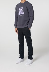 Felpa grigia scura con una grafica di un orso, abbinata a pantaloni cargo neri. Sneakers bianche completano l'outfit. Il materiale sembra morbido e confortevole.