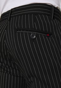 Czarne spodnie w cienkie paski z pojedynczą tylną kieszenią zapinaną na guzik i małą czerwoną metką. Gładka faktura tkaniny.