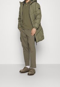 Un manteau long matelassé vert olive, un polo vert olive, un pantalon assorti vert olive, et des chaussures marron clair avec une semelle épaisse. Chaussettes blanches visibles.