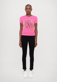 Pielāgots rozā T-krekls ar melnu un zelta "Versace Jeans Couture" grafiku, apvienots ar melniem pieguļošiem biksēm un baltām sporta apaviem.