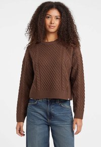 Bruine kabelgebreide trui met een ronde hals en geribde boord, met textuurpatronen en lange mouwen, gedragen met blauwe jeans.
