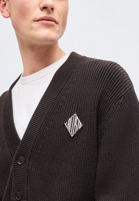 Νεαρό άτομο φοράει καφέ σκούρη πλεκτή ζακέτα με λευκό πουκάμισο από κάτω και μπαλώμα με λογότυπο σε σχήμα διαμαντιού στο στήθος.