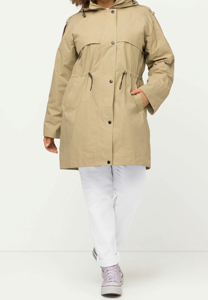 Manteau beige à capuche avec boutons sur le devant et cordon à la taille, porté avec un pantalon blanc et des baskets violet clair, personne debout la main dans la poche.