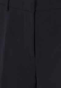 Zbliżenie czarnych spodni materiałowych, ukazujące szczegóły pasa, pętli na pasek i szwu kieszeni bocznej.