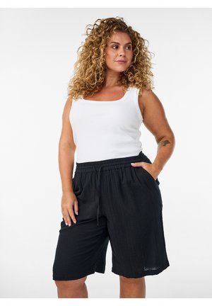 Krøllet-håret kvinde står iført en hvid ærmeløs top og løse sorte knælange shorts med hænderne i lommerne.
