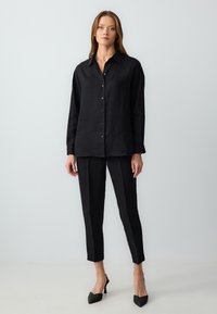 Schwarzes Leinen-Hemd mit langen Ärmeln und Knopfleiste, kombiniert mit schwarzen, eng anliegenden Hosen. Getragen mit spitzen, schwarzen High Heels.