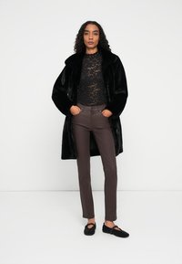 Sort Faux Fur-frakke, blonder langærmet top, brune tætsiddende bukser, sorte vævede slip-on sko; model står mod en hvid baggrund.