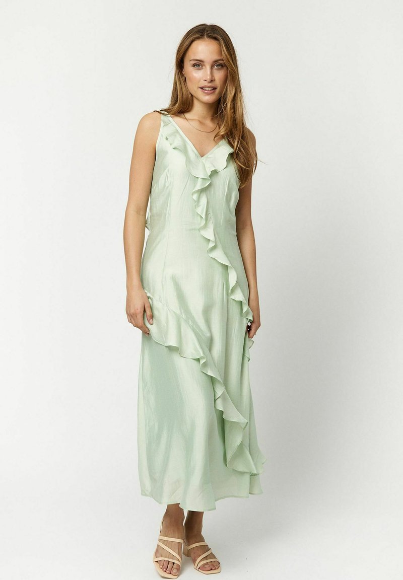 Hellgrünes Satin-Maxi-Kleid mit Rüschendetails an der Front, V-Ausschnitt und Trägern. Kombiniert mit beigen Riemchensandalen.