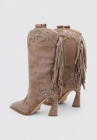Botas de vaquero de ante marrón con puntas afiladas, acentos con flecos, recortes intrincados y adornadas con rhinestones en el talón y la parte superior.