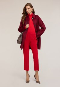 Burgundy ullkappa över en röd polotröja, i kombination med röda skräddarsydda byxor och bruna klackskor. Modellen håller en grå handväska med en kedjestropp.