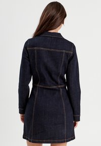 Vestido de mezclilla en azul oscuro con mangas largas, una cintura ceñida con cinturón y costuras de contraste. Presenta un cuello de camisa y detalle de yugo en la parte posterior.