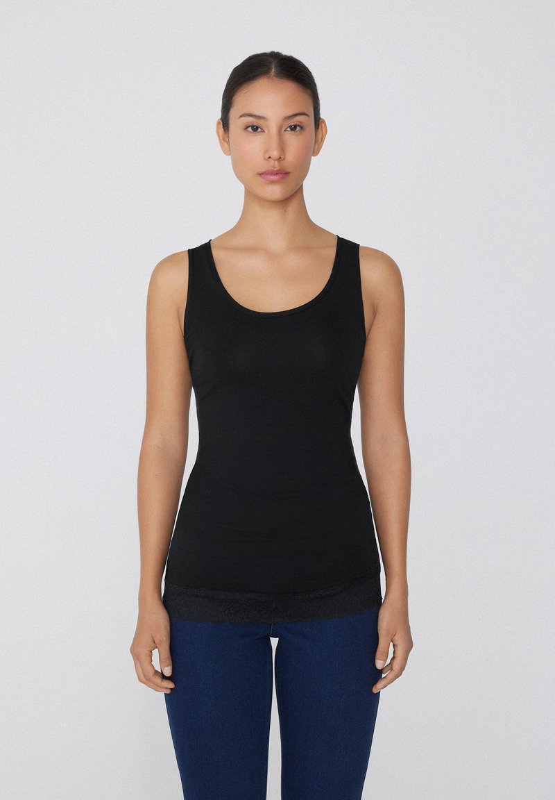 Schwarzes Tanktop aus glattem Stoff mit einem Rundhalsausschnitt und einer Spitzenborte am Saum. Kombiniert mit dunkelblauen Jeans.