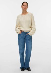 Kremowy sweter w warkoczowy splot z szerokimi rękawami w połączeniu z jasnoniebieskimi spodniami typu straight-leg. Czarne płaskie buty dopełniają outfit. Proste tło.