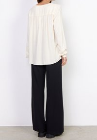 Blouse crème à manches longues avec un col rond et un détail froncé au dos. Assortie à un pantalon noir à taille haute et à des chaussures noires.