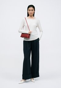 Crèmekleurig long-sleeve top, donkere wijde broek met manchetten en een rode schoudertas. Model draagt witte instap-schoenen, met minimaal textuur overal.