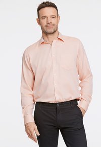 Camisa de botón de un suave tono rosa claro con un bolsillo en el pecho y un cuello interior estampado, combinada con pantalones negros de corte relajado y textura suave.